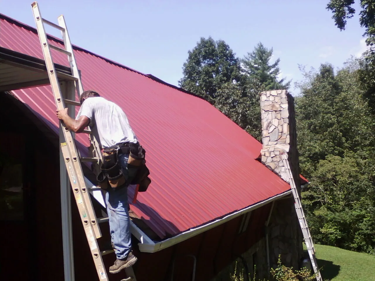 Roofer on a ladder beside a red metal roof and stone chimney — Soffit & Fascia Repair by Roofing Brothers in Telford